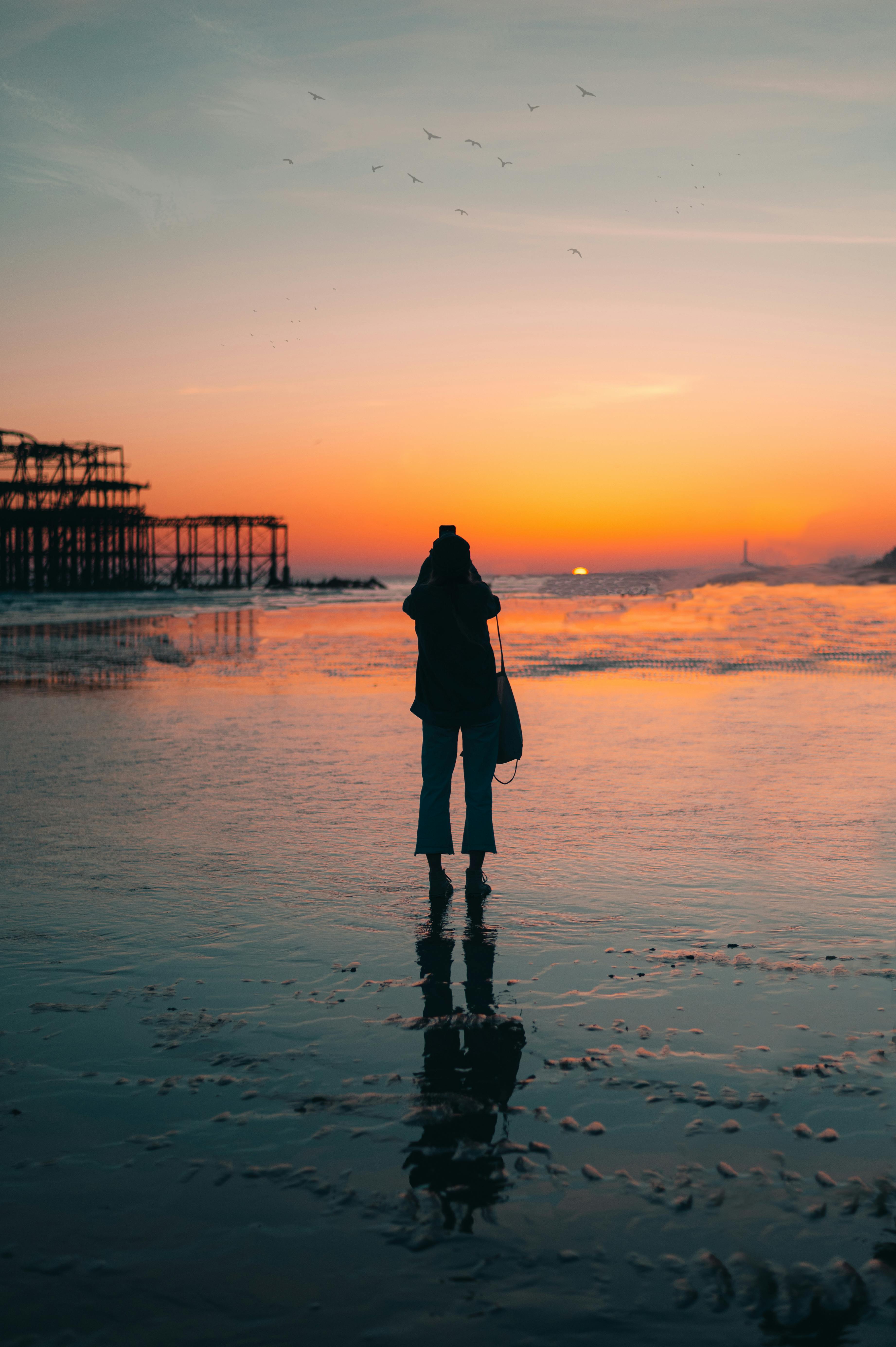 Silhouetted figure of a woman on the beach at sunset with a pier and dramatic sky.