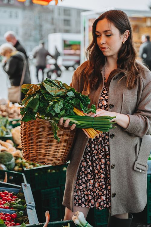 Free A woman examines fresh greens at an outdoor market, holding a woven basket. Stock Photo