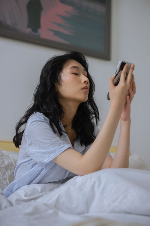 Free Young Asian woman sitting on bed using her smartphone at home, looking relaxed and comfortable. Stock Photo