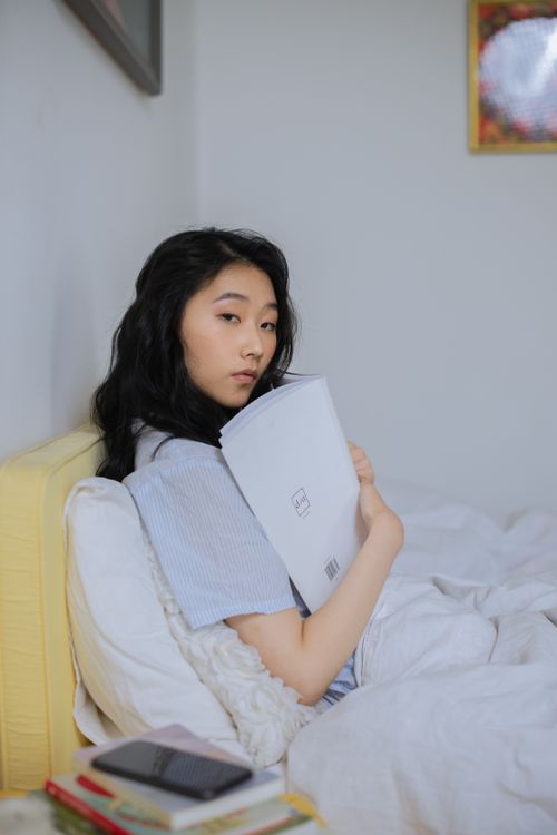 Free Asian woman relaxing in a comfy bedroom, enjoying reading a book while sitting on a bed. Stock Photo
