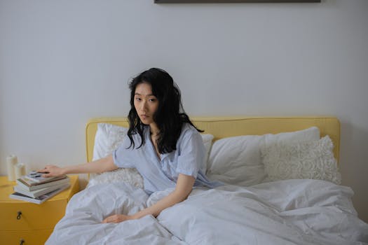 Asian woman sitting in a cozy bedroom with books and white linens, creating a serene ambiance.