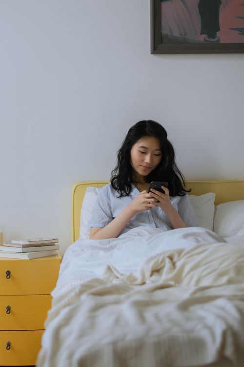 Free Young woman relaxing in bed, looking at her smartphone in a cozy bedroom. Relaxed and content atmosphere. Stock Photo