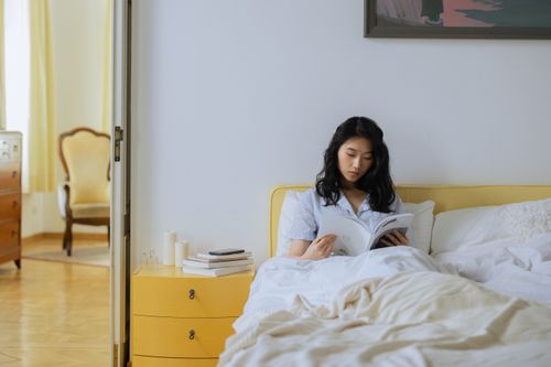 Free Asian woman relaxing and reading a book in a bright, cozy bedroom setting. Stock Photo