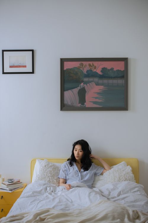 Free Asian woman sitting on bed in a minimalist bedroom, relaxing in a serene indoor setting. Stock Photo