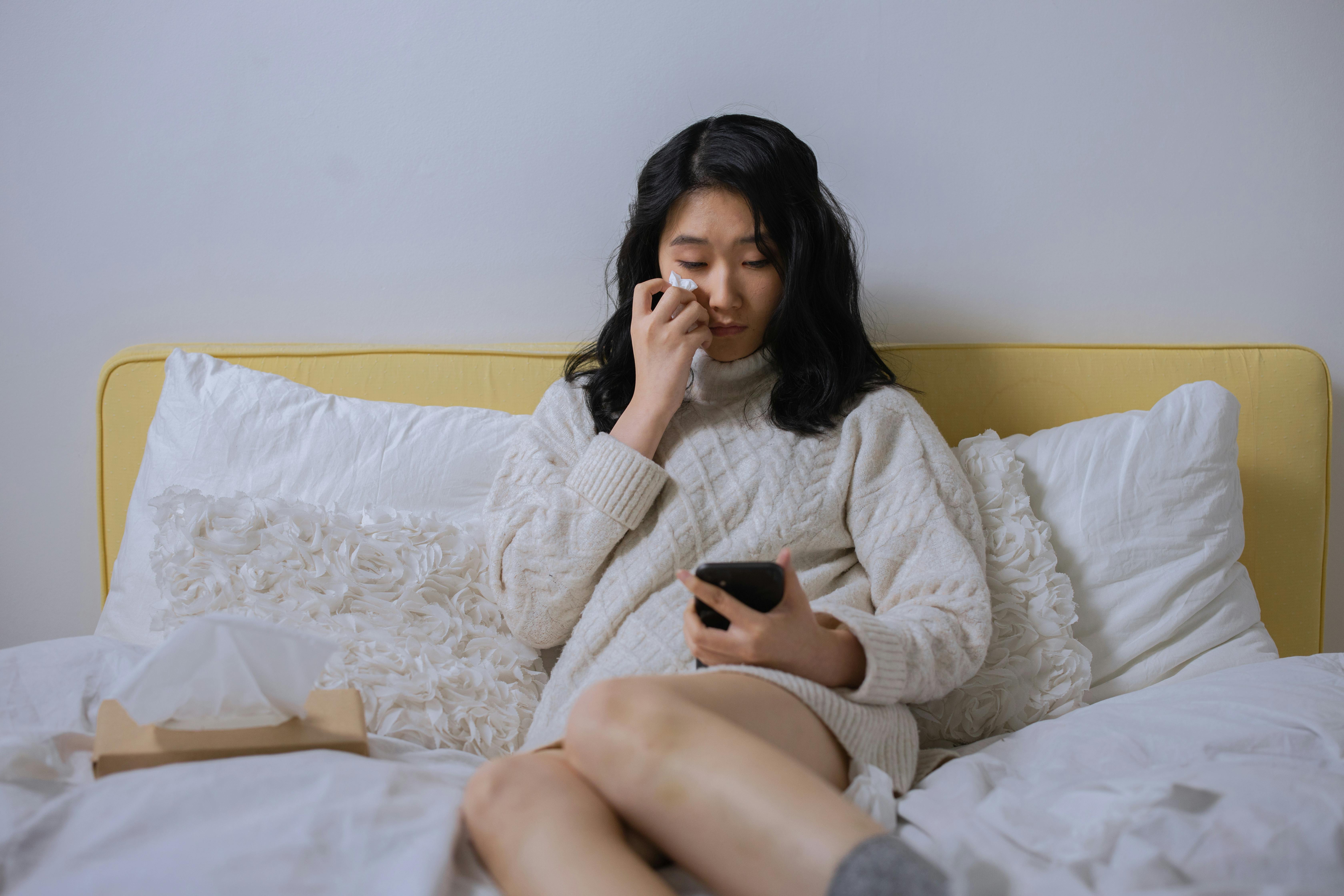 a sad woman looking at her cellphone while sitting on a bed