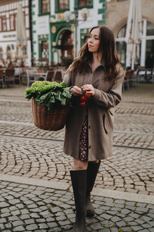 Free A woman in a brown coat carries fresh vegetables in a basket, walking in a quaint European square. Stock Photo