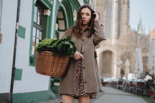 Woman in Brown Coat Carrying Basket Full of Green Vegetables