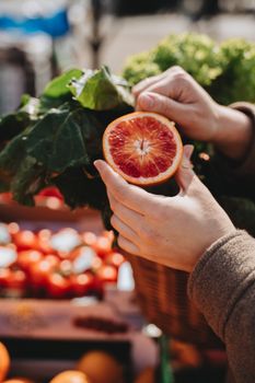 Close-up of hands holding a sliced orange with vegetables in a market setting, showcasing freshness and vibrancy.
