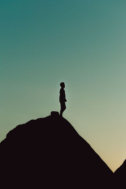 A lone figure stands silhouetted on a mountain peak, backlit by the warm, fading hues of sunset.