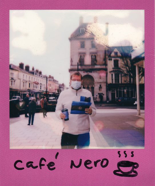 Polaroid photo of a man wearing a face mask holding coffee outdoors in a city.