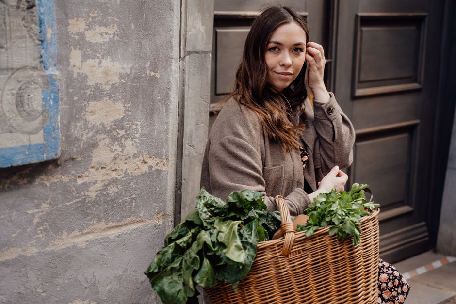 A woman smiling while holding a wicker basket filled with fresh green vegetables outdoors.