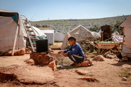 Free A young boy in a rural Idlib camp tends to a cooking pot amidst tents and rugged terrain. Stock Photo