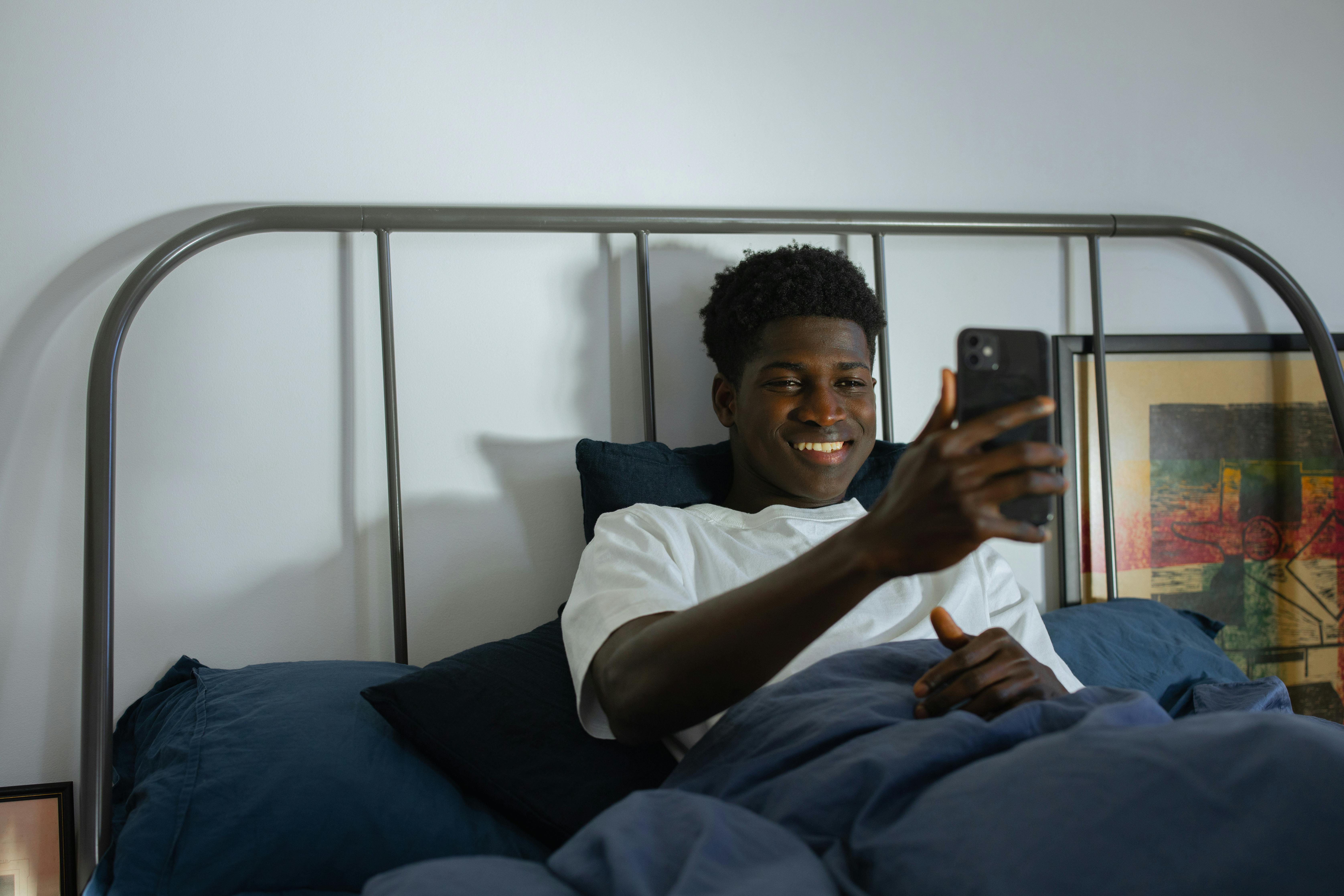 A man smiling while using a smartphone in a modern bedroom setting, illustrating relaxation and connectivity.