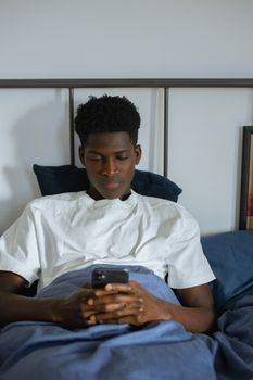 A young man in a white shirt using a smartphone while resting in bed, depicting modern lifestyle.