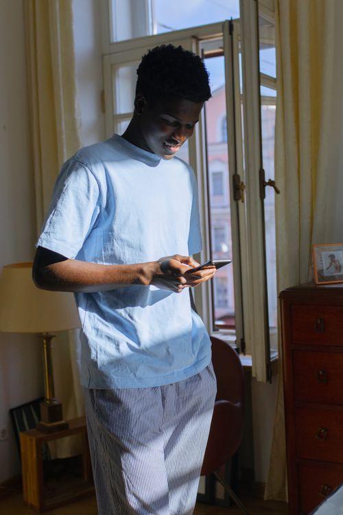 Free A young man in a blue shirt and striped pants checks his smartphone indoors by the light of a window, smiling. Stock Photo