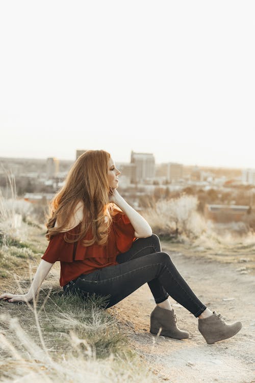 Free Side view of stylish young woman in blouse and boots sitting on pathway while looking away in town Stock Photo