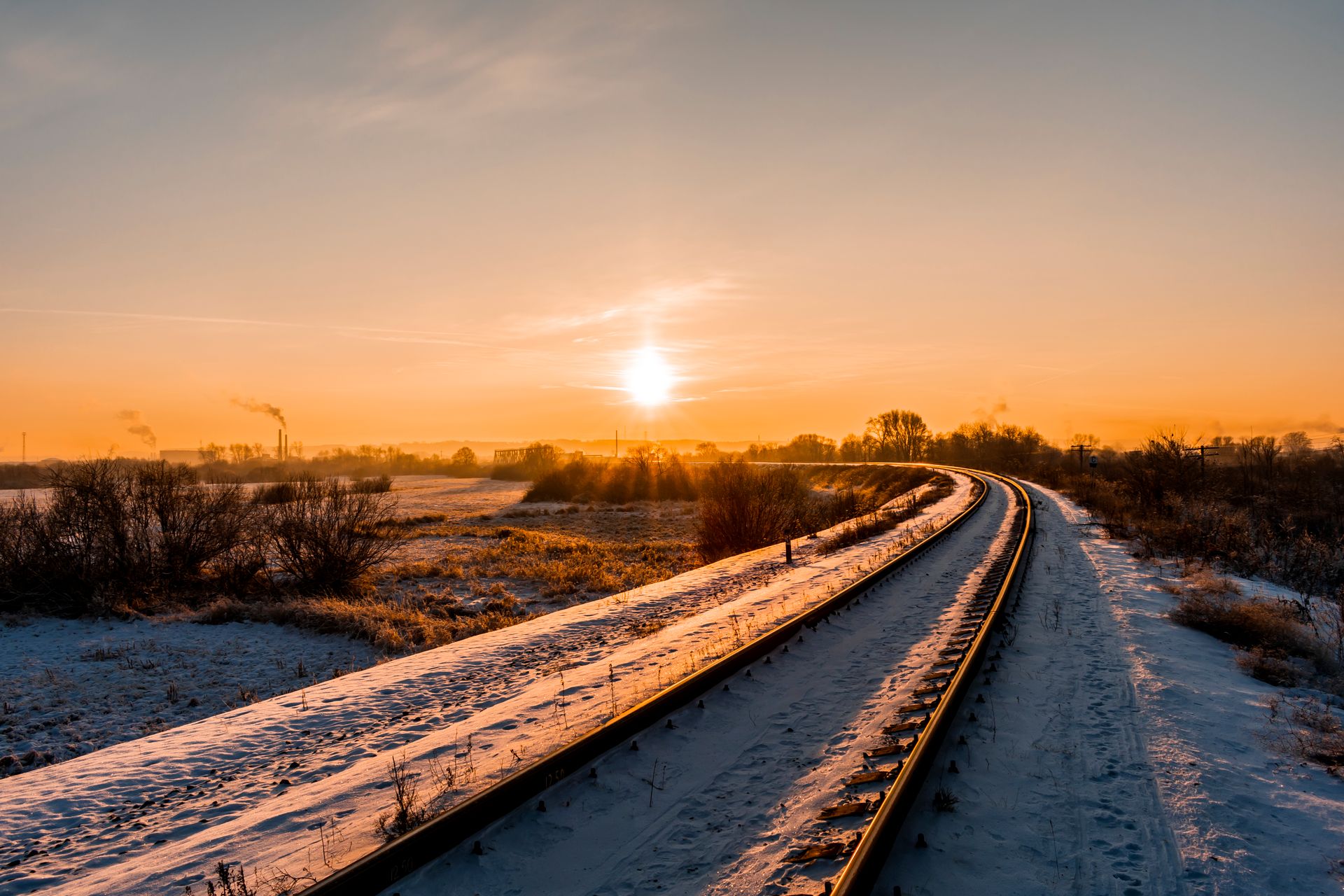 A serene winter sunrise casts a warm glow over snowy railroad tracks in the countryside.