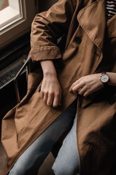 High angle of crop unrecognizable female wearing stylish coat and jeans with wristwatch while sitting in chair near window in bright room