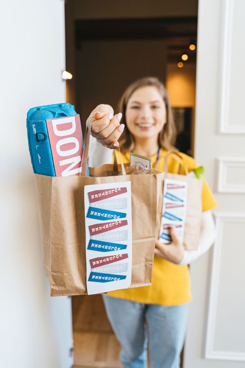 Free Happy woman delivering groceries wearing yellow shirt, standing in the doorway holding paper bags. Stock Photo