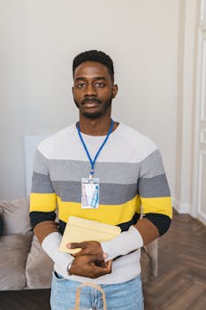 A young man with short hair confidently poses indoors holding a folder.