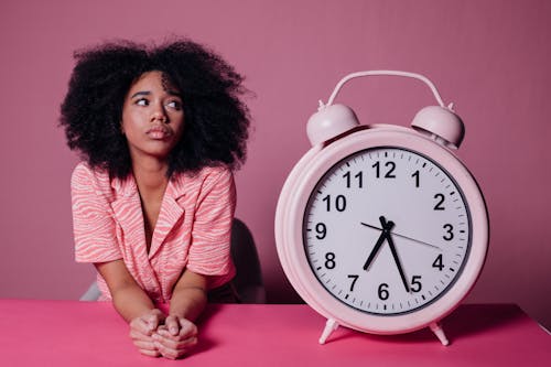 Woman in Pink Long Sleeve Shirt Sitting Beside White and Pink Alarm Clock