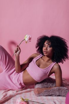 Elegant woman with afro hair in stylish pink outfit holding a rose against a pink background.