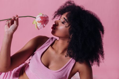 Free A woman with afro hair in a pink crop top smelling a pink rose against a matching background. Stock Photo