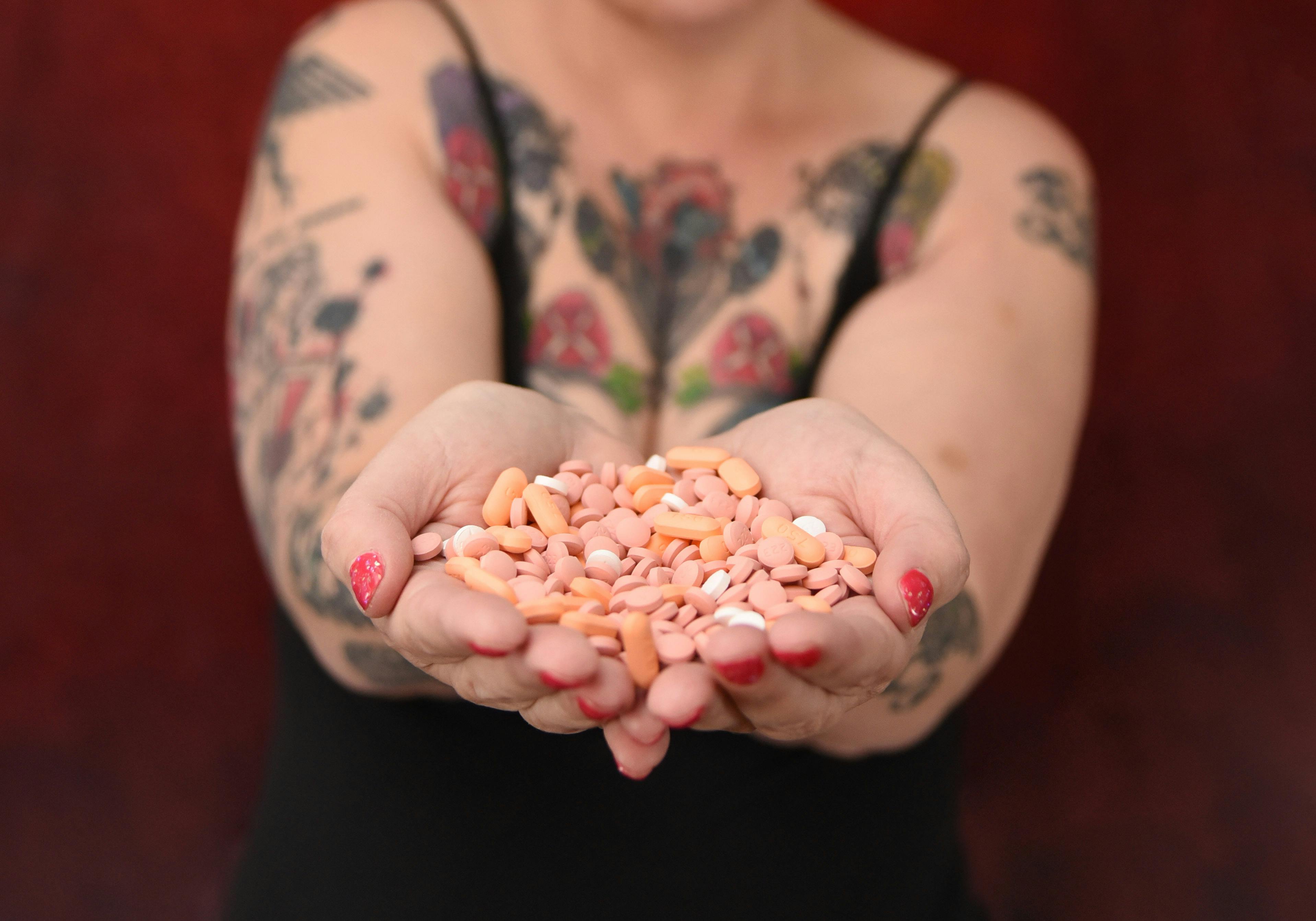 Close-up of tattooed woman offering a pile of colorful pills with outstretched hands.