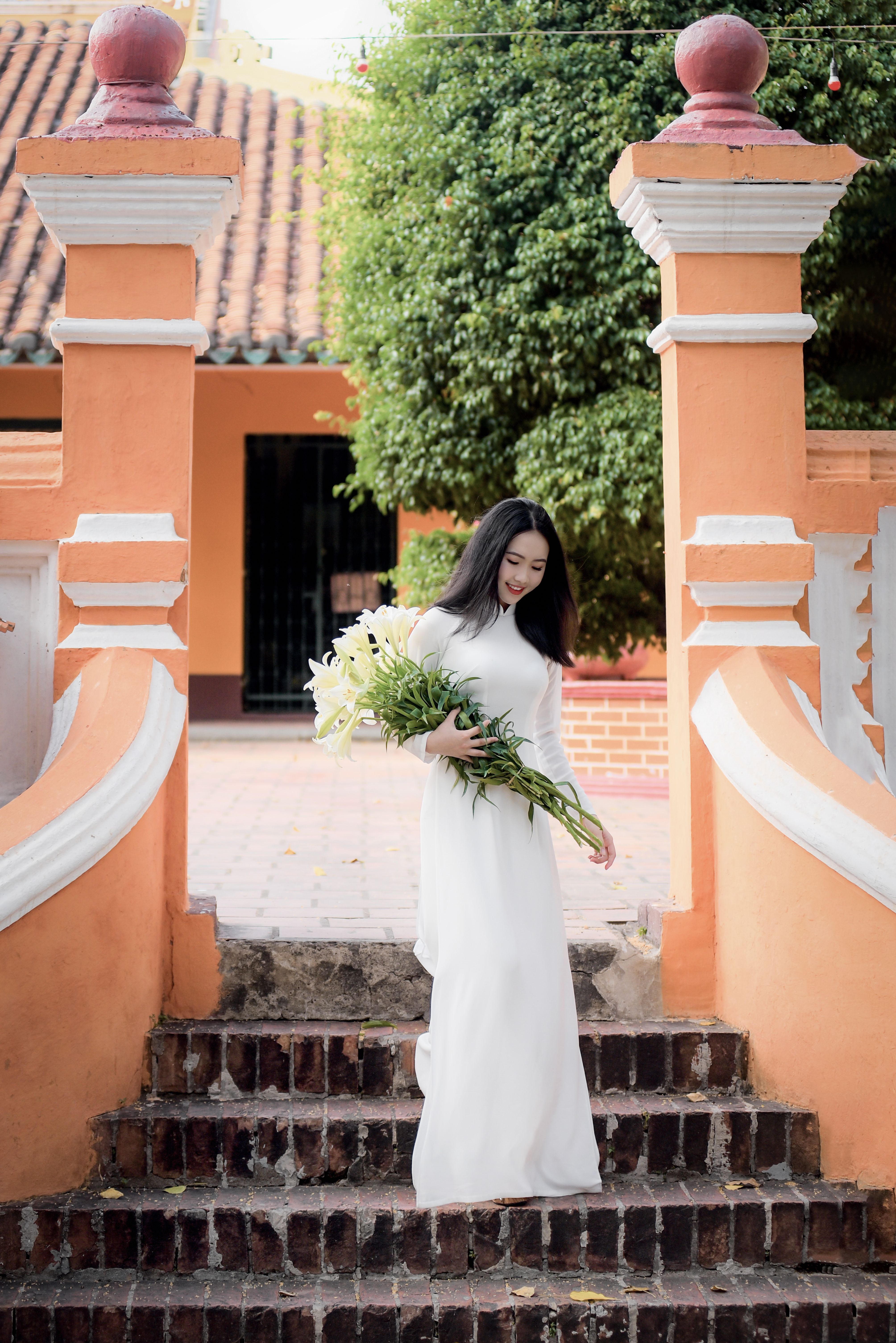 Beautiful Asian woman in white Ao Dai holds flowers while descending brick stairs with traditional architecture backgrou