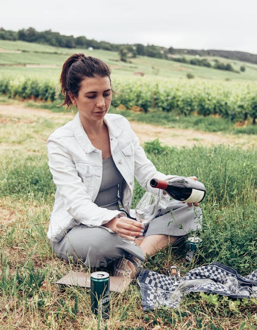 Free A Woman in White Jacket Sitting on Grass Holding a Wine Bottle Stock Photo