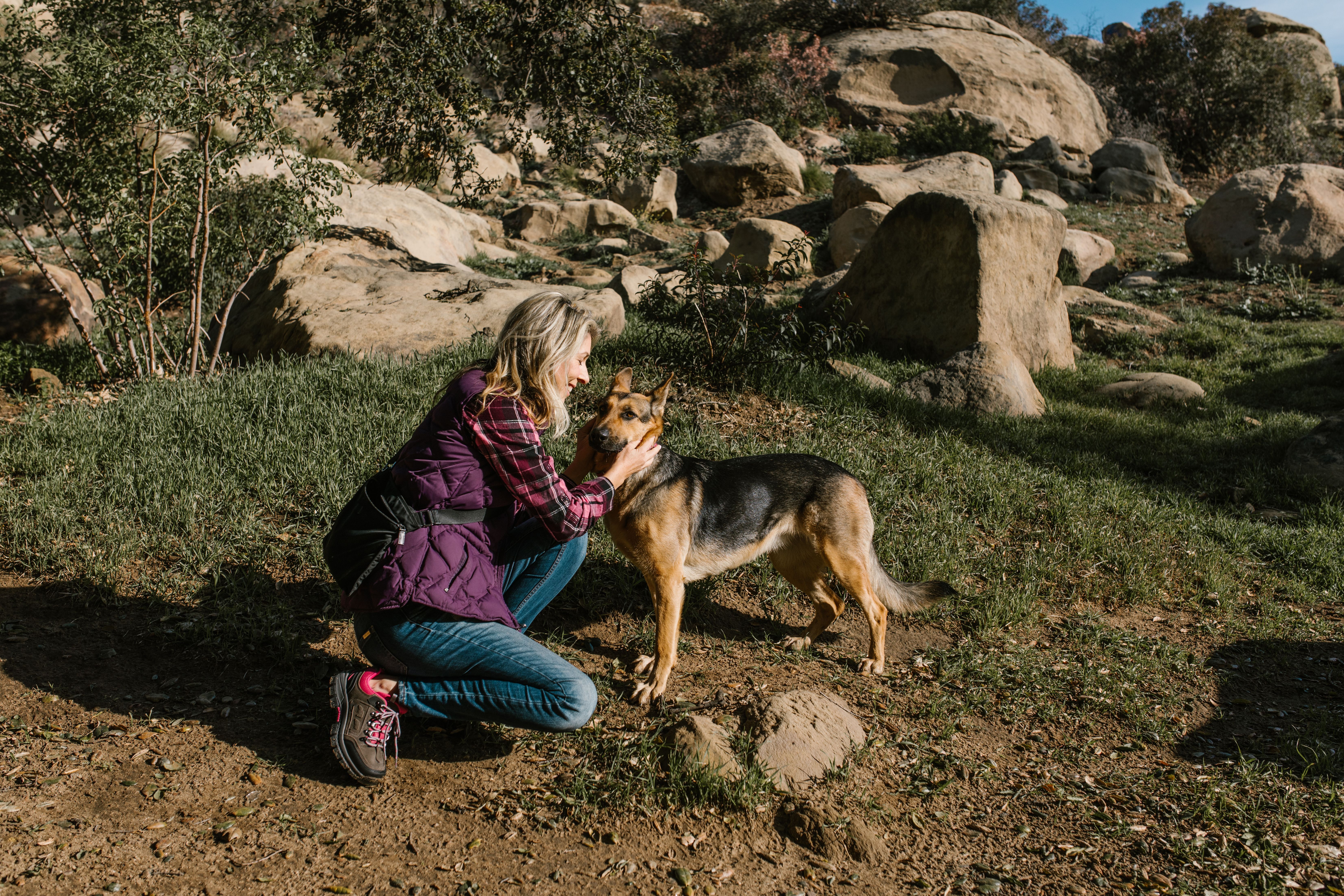 A woman kneels to pet her dog in a rocky, sunny outdoor landscape.