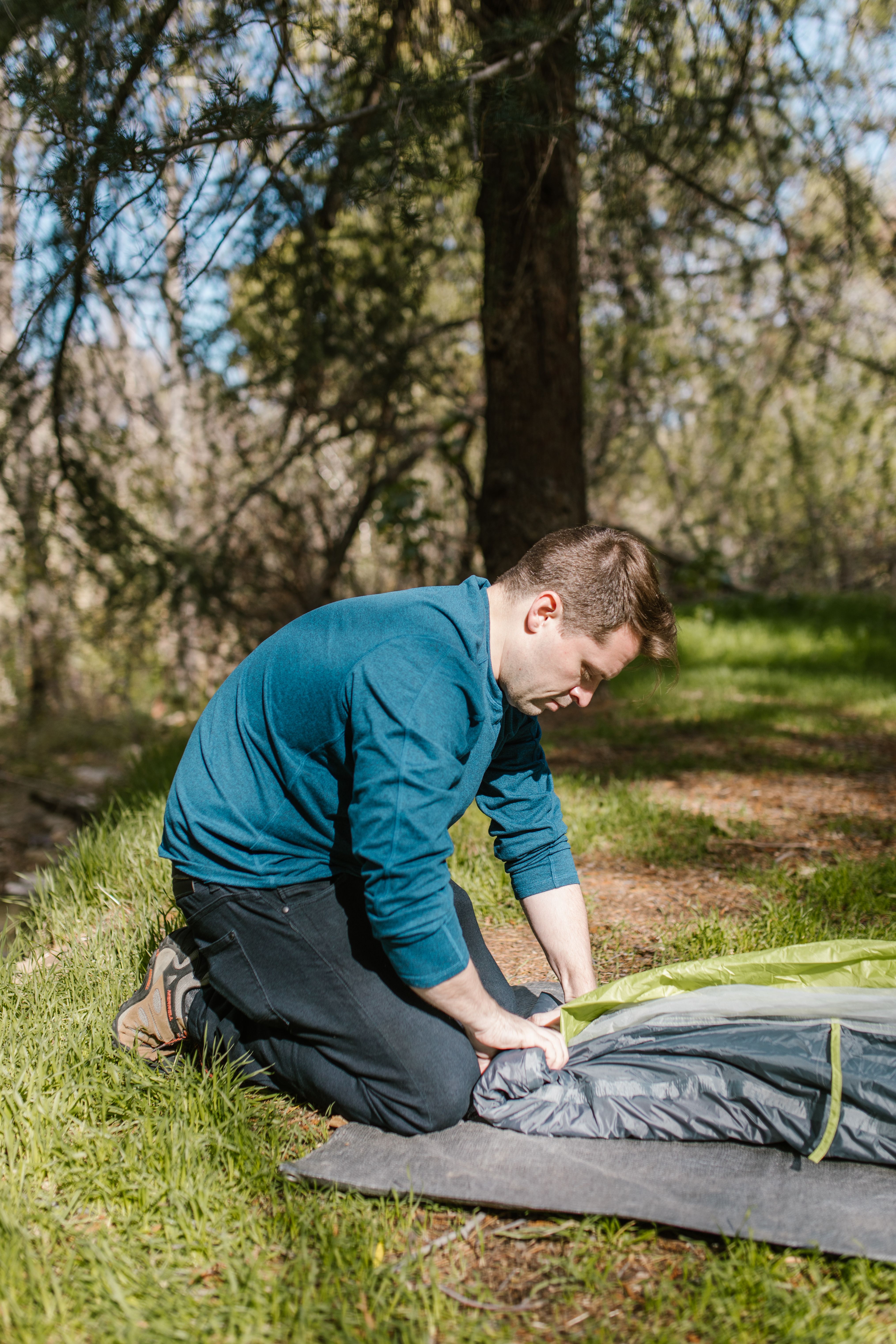 A man in a blue shirt kneels on grass setting up a tent in a forest during the day.