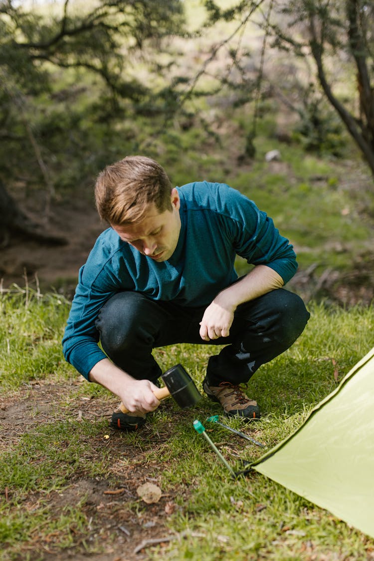 A Man Wearing Blue Long Sleeves Shirt Hammering A Tent Peg