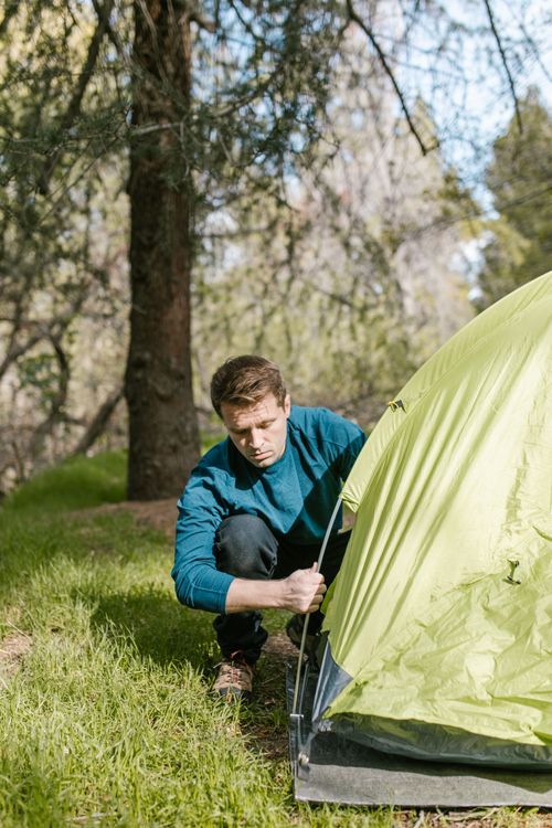 Free Man wearing blue sweater sets up a green tent in a sunny forest clearing. Stock Photo