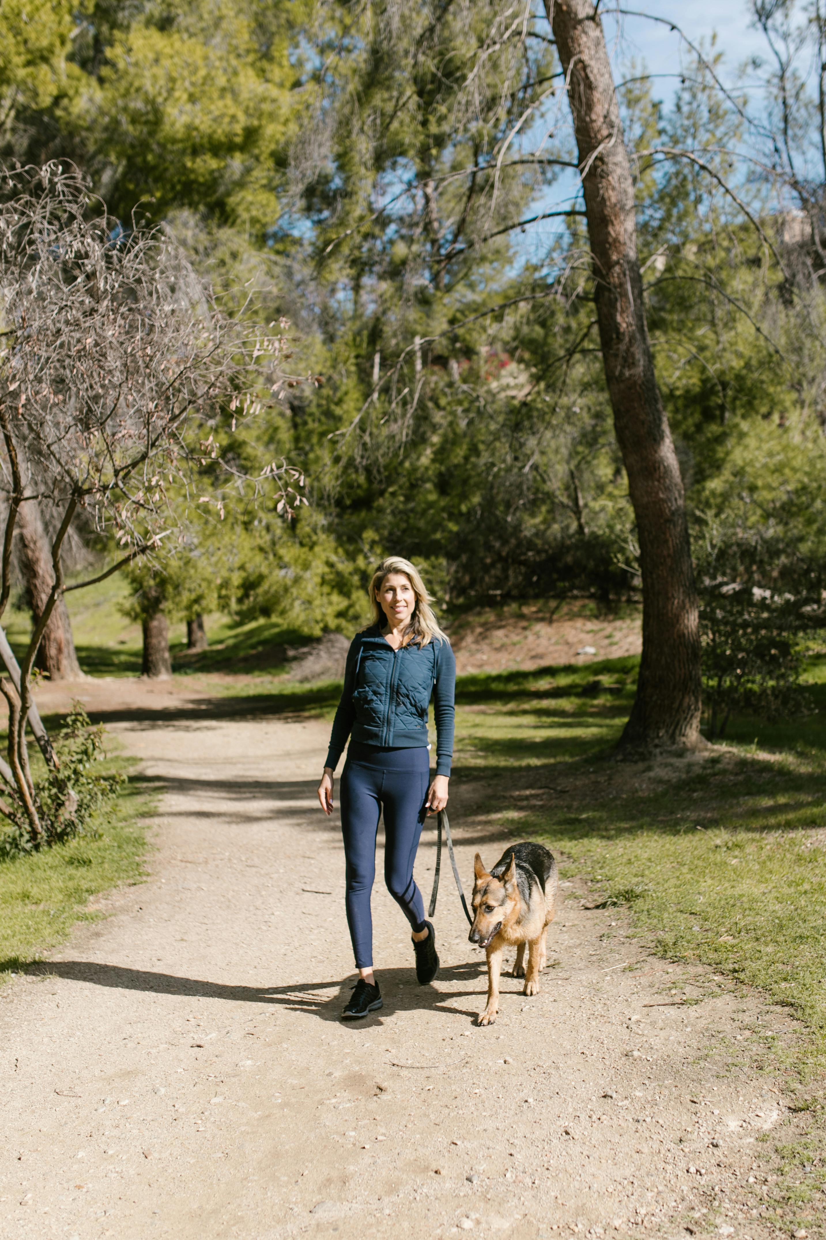 A woman walks her German Shepherd in a scenic forest trail during springtime.