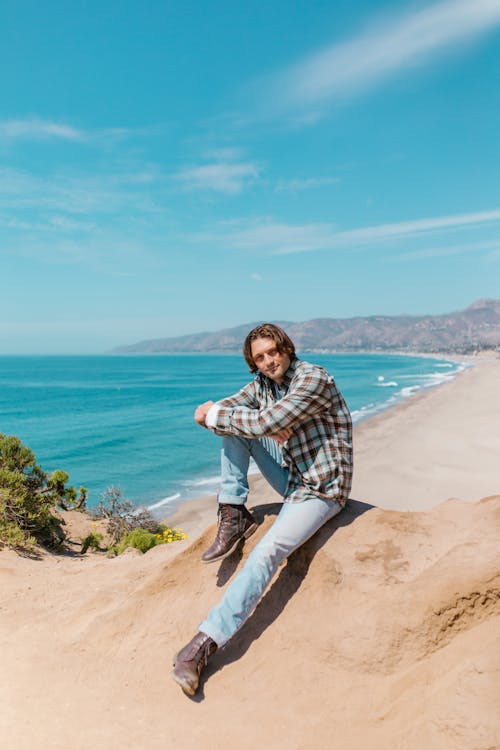 Man in Plaid Long Sleeves and Denim Jeans Sitting on Rock Near Body of Water