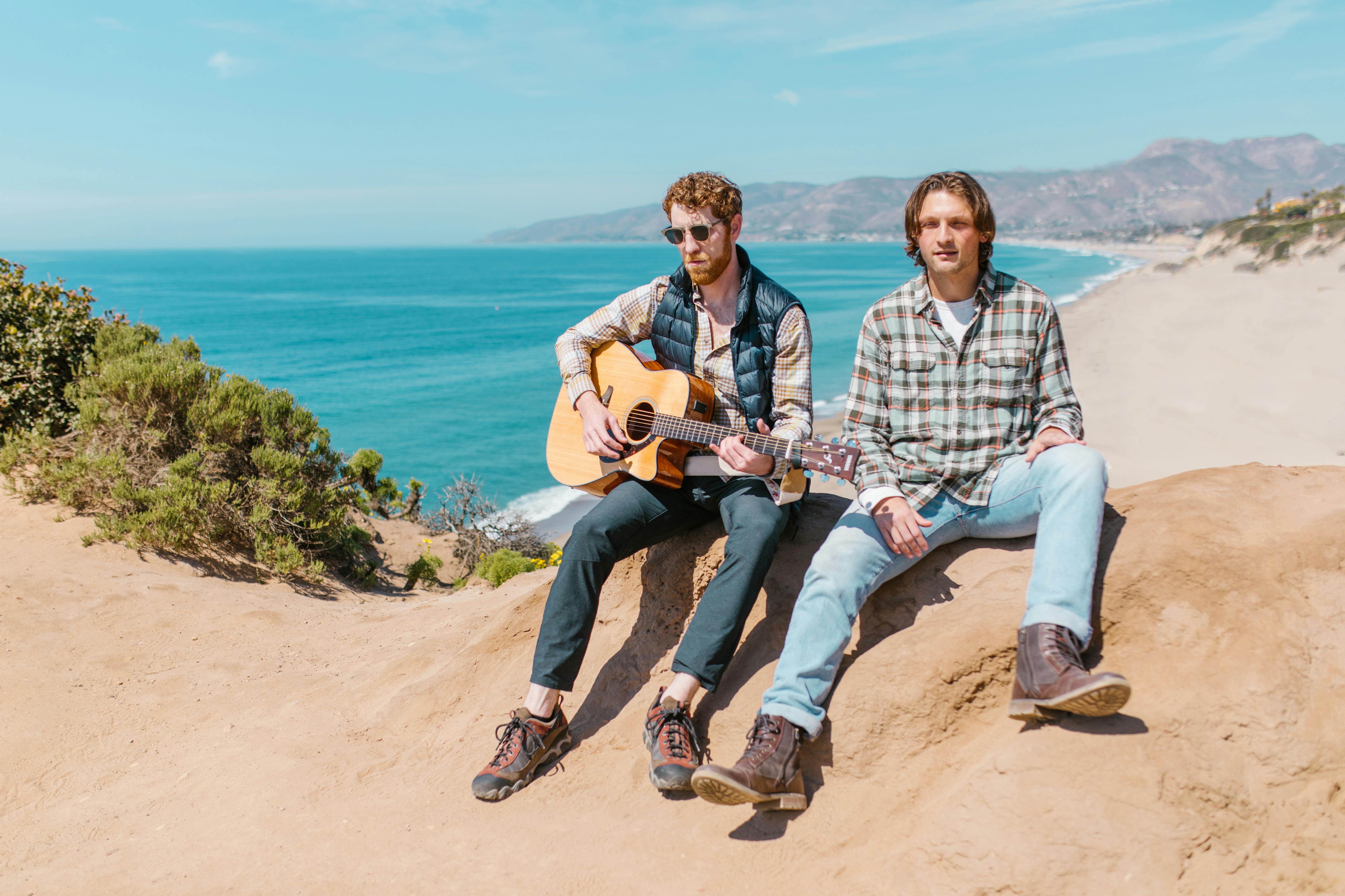 Two men with a guitar relaxing by the beach, capturing a carefree and adventurous spirit.