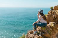 戴着帽子的女人坐在悬崖上，在晴朗的天空下欣赏海景