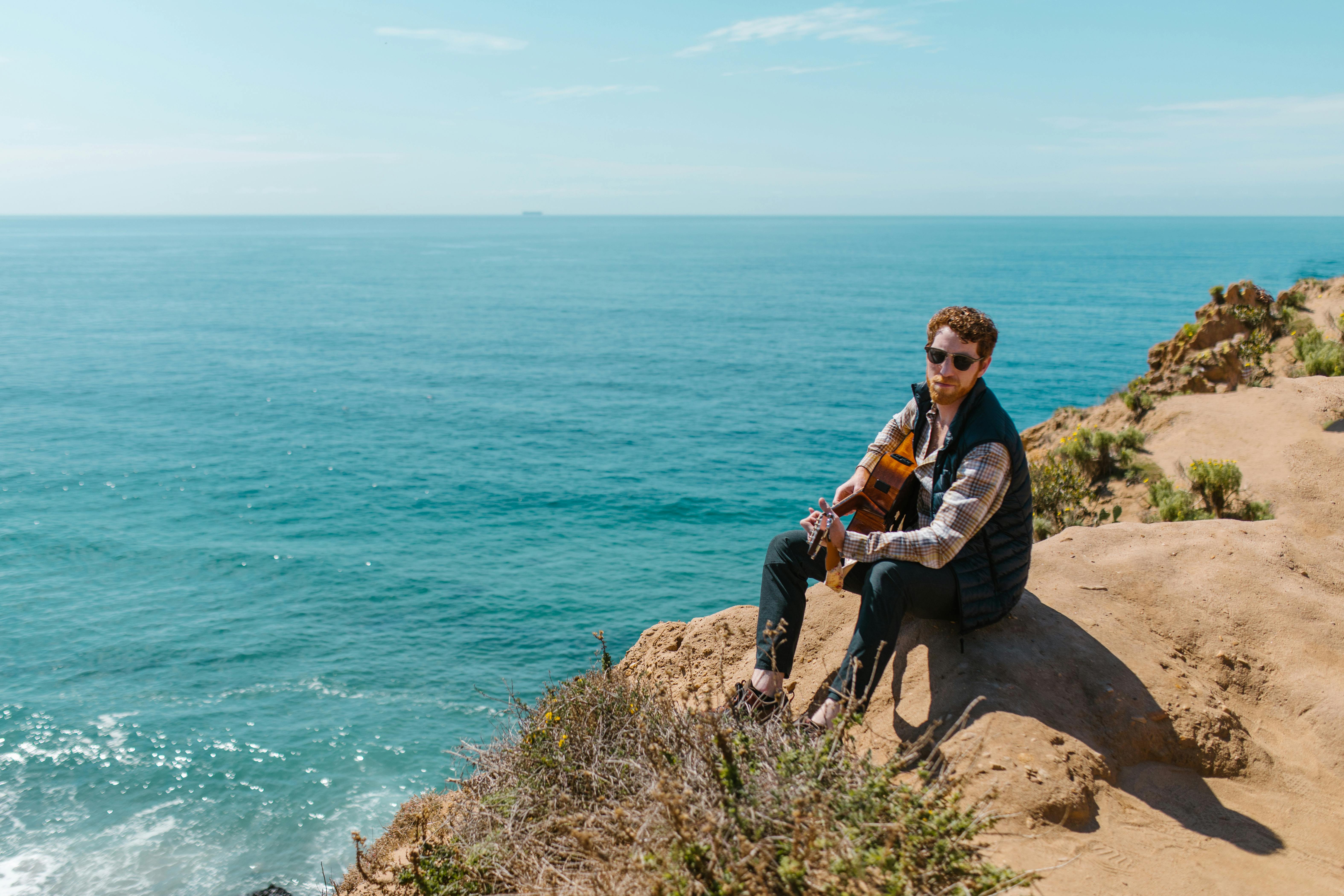 A man in a plaid shirt plays guitar on a coastal cliff, overlooking the serene sea.