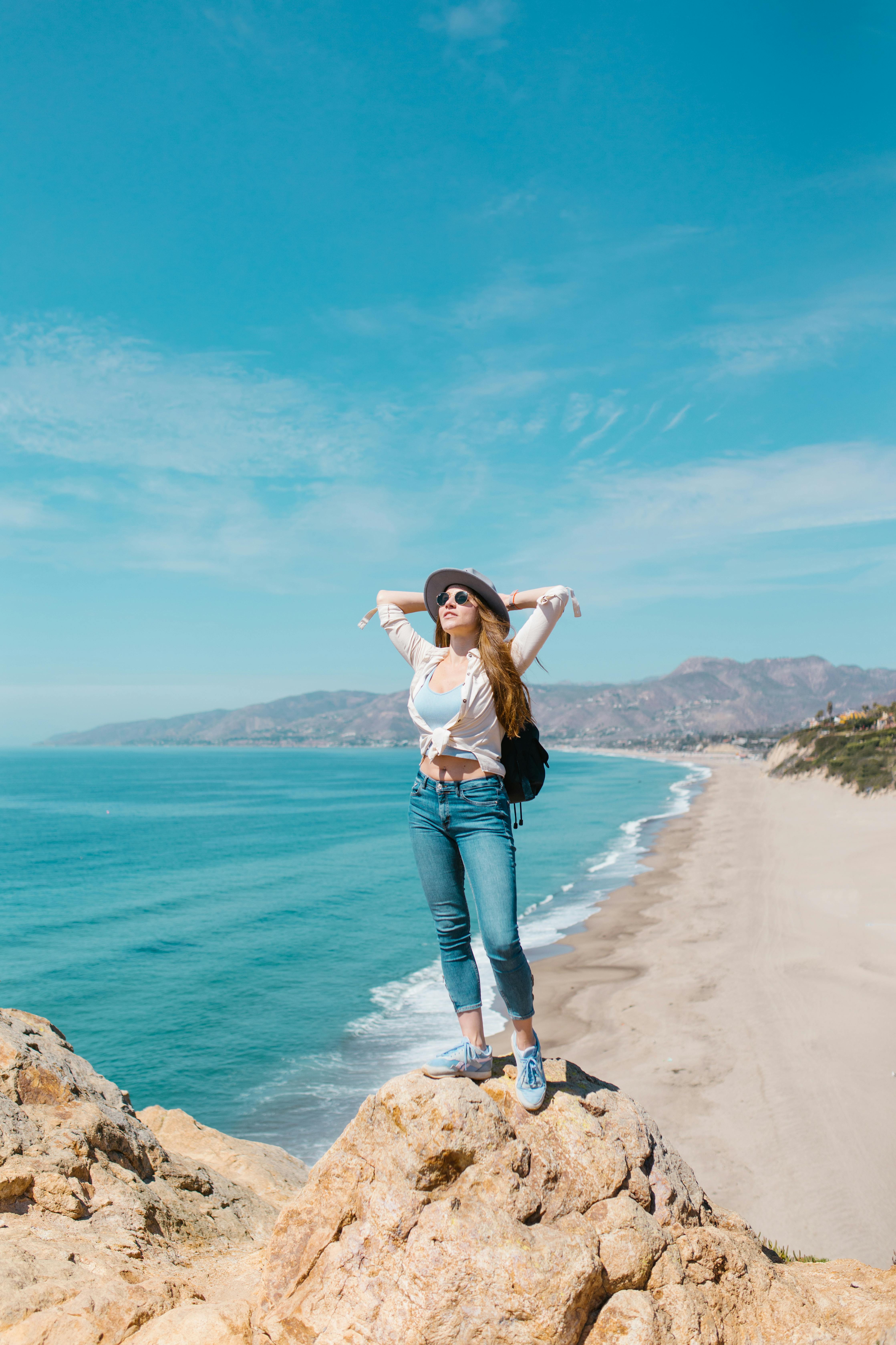 woman in white shirt and blue denim jeans standing on rock formation by the sea during