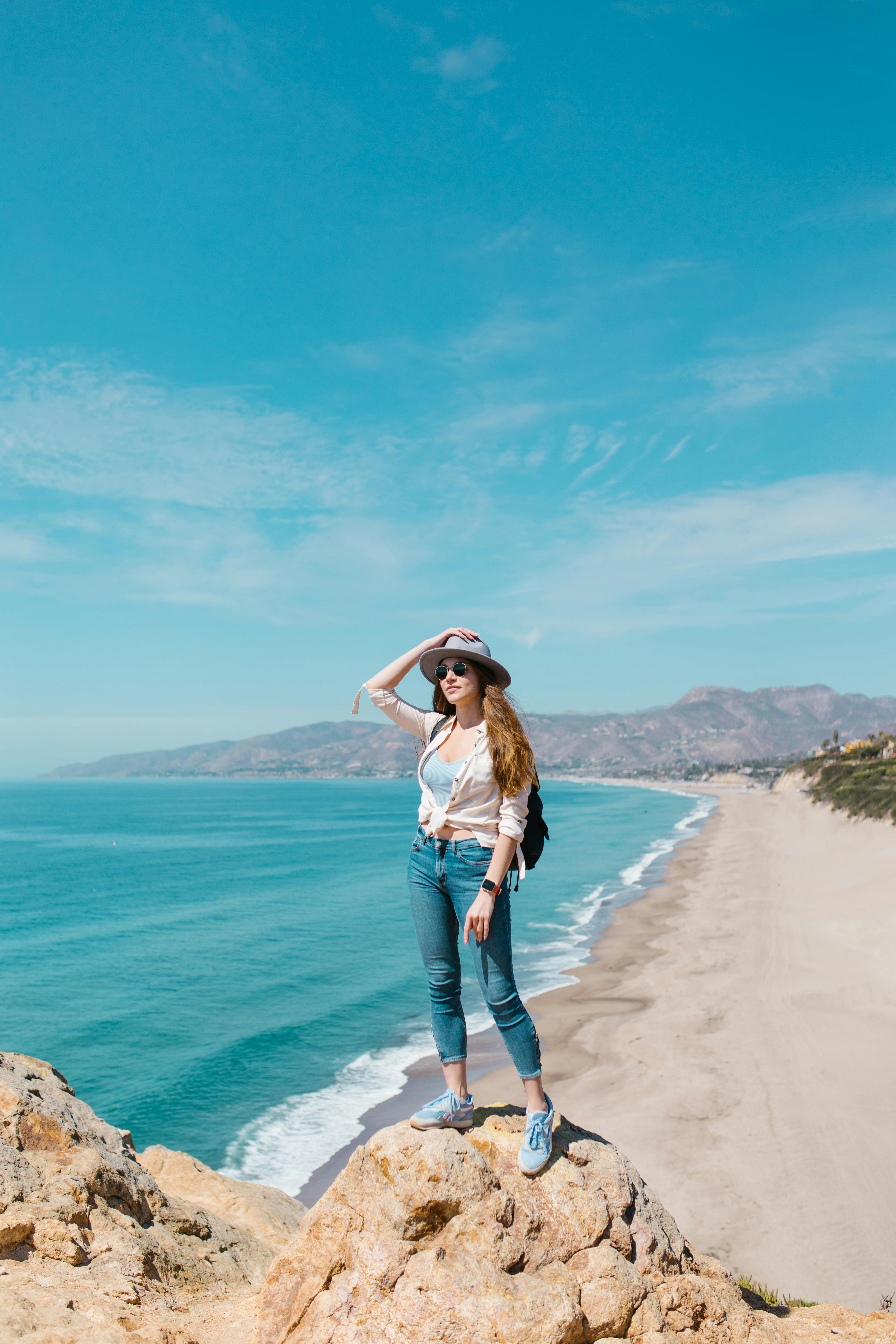 woman in white shirt and blue denim jeans standing on brown rock formation by the sea