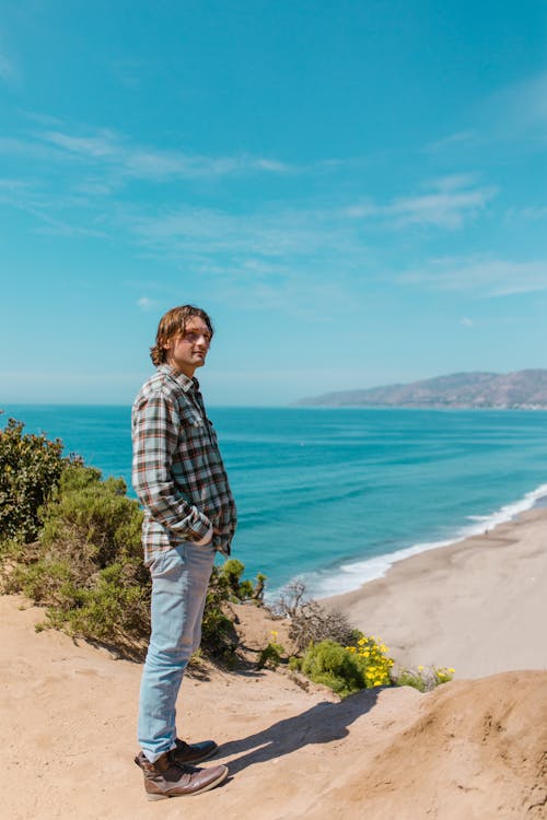 Man in Checkered Long Sleeves Standing on the Beach under the Blue Sky