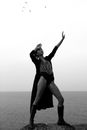 Black and white of young woman in bodysuit and boots touching forehead while looking up with raised arm against ocean
