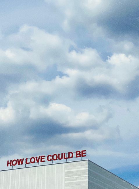 Building facade in Bochum, Germany with 'HOW LOVE COULD BE' sign against a cloudy sky.