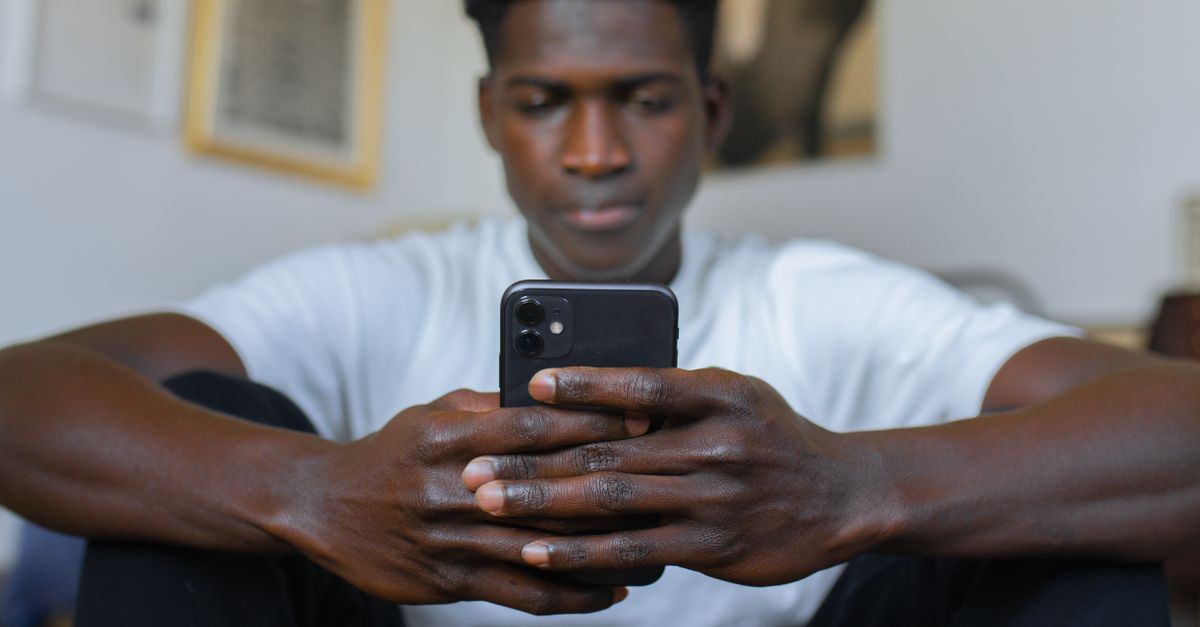 Um homem em uma camisa branca está usando seu smartphone com atenção enquanto está sentado dentro de casa.