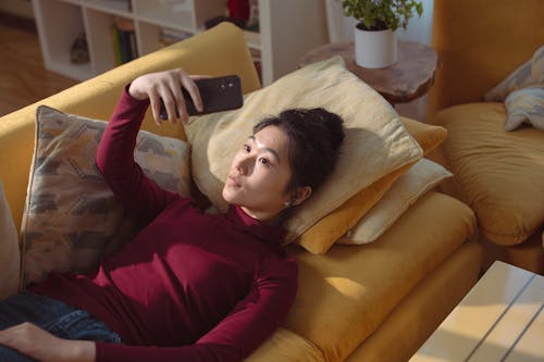Free High-Angle Shot of a Woman in Maroon Sweater Lying on Sofa while Taking Photo of Herself Using a Cellphone Stock Photo