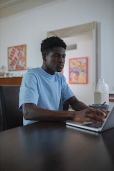 Focused young man working on a laptop at home with a beverage nearby. Ideal for lifestyle and remote work themes.