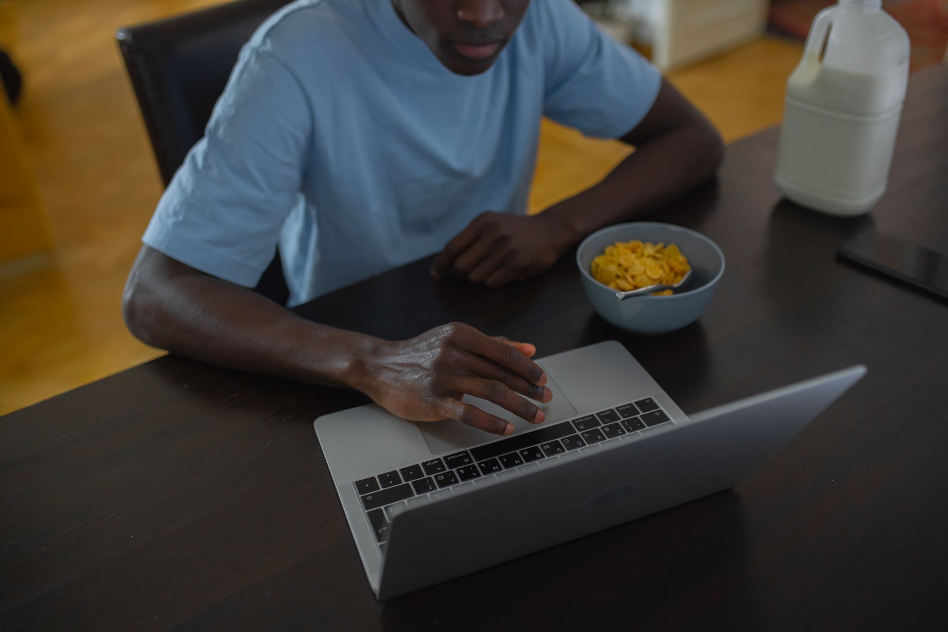 A man in casual clothing using a laptop at home, with cereal and milk on the table.