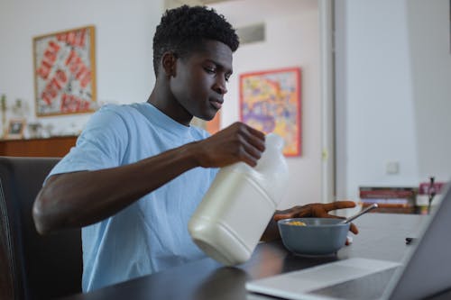 Teenage Boy Sitting at the Table and Pouring Milk into a Bowl with Cereal 