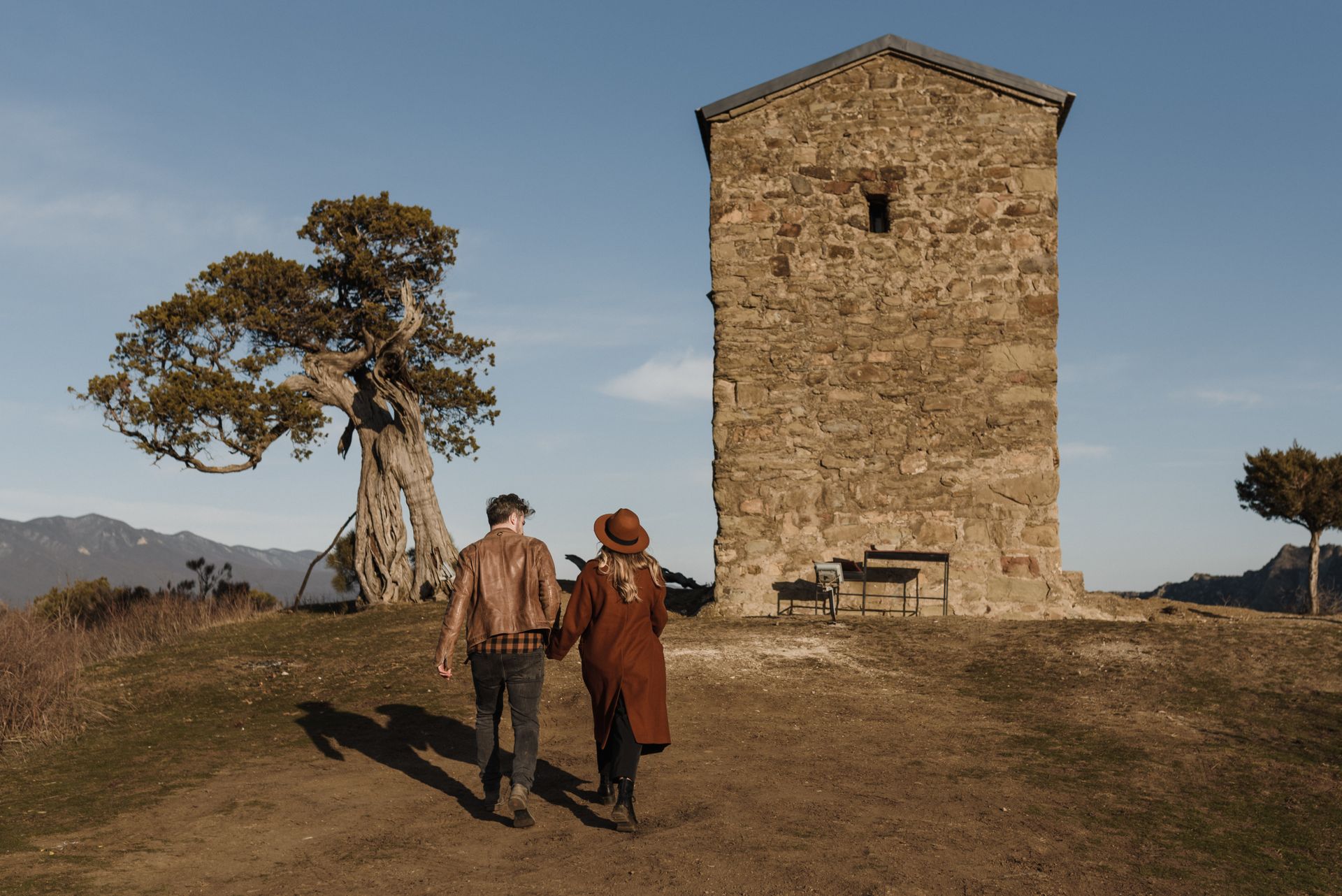 A couple strolls hand in hand towards a rustic stone tower in a serene rural setting.